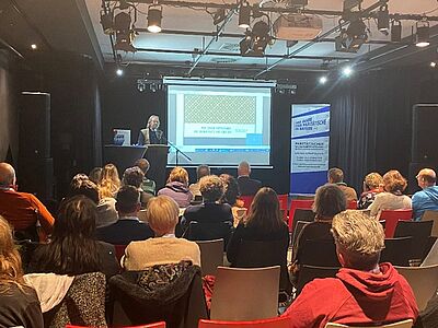 Foto von der Auftaktveranstaltung: Ein Vortragender steht an einem Rednerpult vor einer Leinwand, auf der eine Präsentationsfolie angezeigt wird. Das Publikum sitzt in mehreren Reihen in einem Veranstaltungsraum und hört zu.