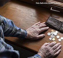 Ein älterer Mann stützt sich auf ein Fensterbrett. Vor seinen Händen liegen eine Geldbörse und kleine und größere Münzen.
