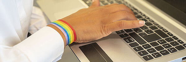 Hand einer Frau auf eine Laptop-Tastatur. Um das Handgelenk trägt die Frau ein Armband in Regenbogenfarben.
