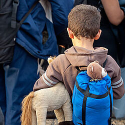 Ein kleiner Junge steht in einer Gruppe von Menschen. Er hat einen Rucksack auf, aus dem ein Teddy rausschaut.