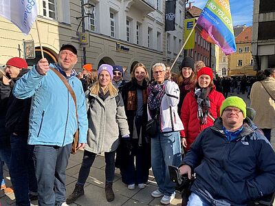 Eine Gruppe von acht Menschen steht bei sonnigem Wetter in der Regensburger Innenstadt und lächelt in die Kamera. Einige halten bunte Fahnen mit Botschaften für Vielfalt und Toleranz. Die Personen tragen Winterkleidung, darunter Jacken, Mützen und Schals. Im Hintergrund sind weitere Demonstrierende und Häuserfassaden zu sehen.
