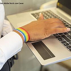 Hand einer Frau auf eine Laptop-Tastatur. Um das Handgelenk trägt die Frau ein Armband in Regenbogenfarben.