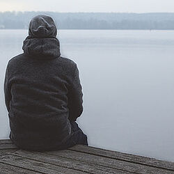 Ein Mann sitzt allein auf einem Steg am See und schaut auf das Wasser.